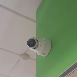 Kamera przemysłowa kopułkowa zamontowana na zielonej ścianie w rogu pomieszczenia, widoczny biały sufit podwieszany i fragment drewnopodobnej listwy przypodłogowej.