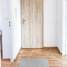 Jasne wnętrze przedpokoju z drewnianą podłogą i beżową wycieraczką, widoczne drzwi wejściowe z drewnopodobnym wzorem i metalową klamką z kluczami, białe ściany i ościeżnice drzwi.