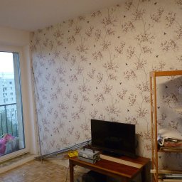 Sławek Maciążek - Pokój z tapetą w drobne kwiaty i ptaki, widoczny fragment balkonu z suszarką na pranie, na podłodze panele, częściowo odsłonięta ściana, niski stolik z telewizorem i skrzynką narzędziową.