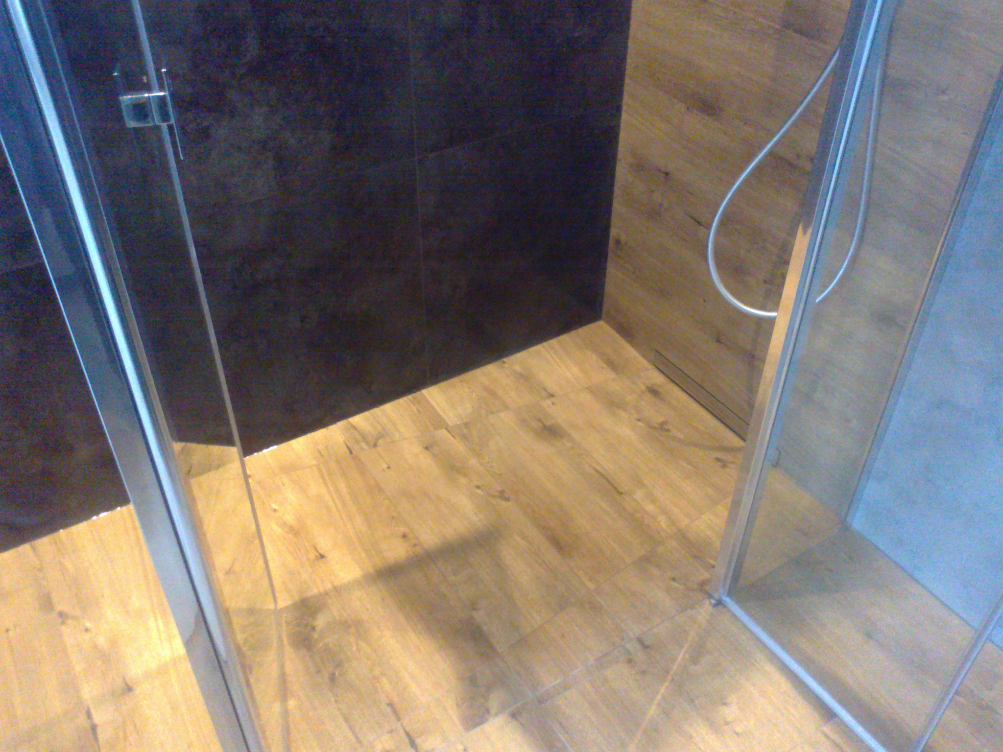 Nowoczesna kabina prysznicowa z drewnopodobnymi płytkami na podłodze i ciemnymi płytkami na ścianie, widok z góry na narożnik.