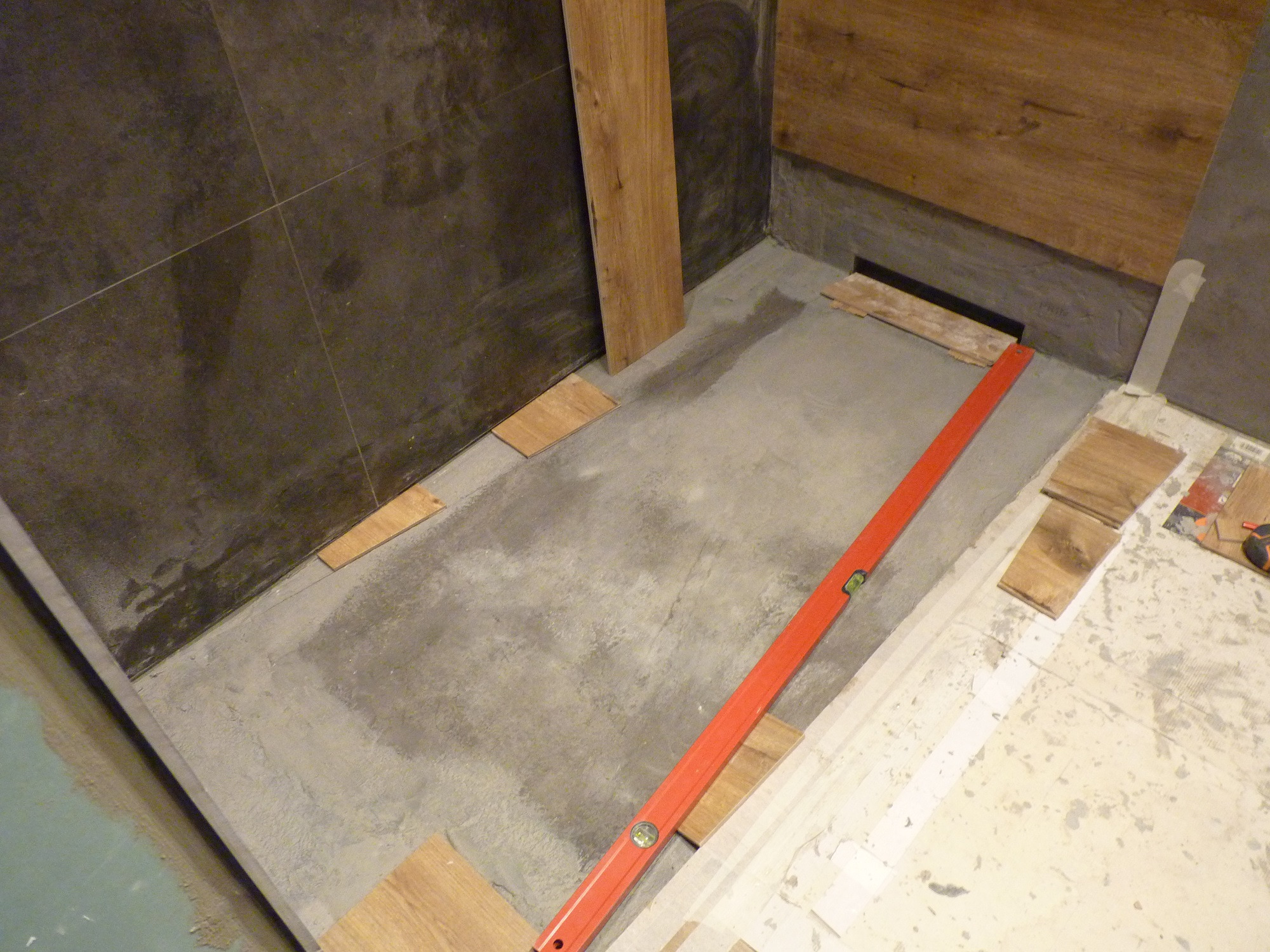 Przygotowanie podłoża pod drewnopodobne panele w łazience: wylewka, poziomica, fragmenty paneli i płytki na ścianie.