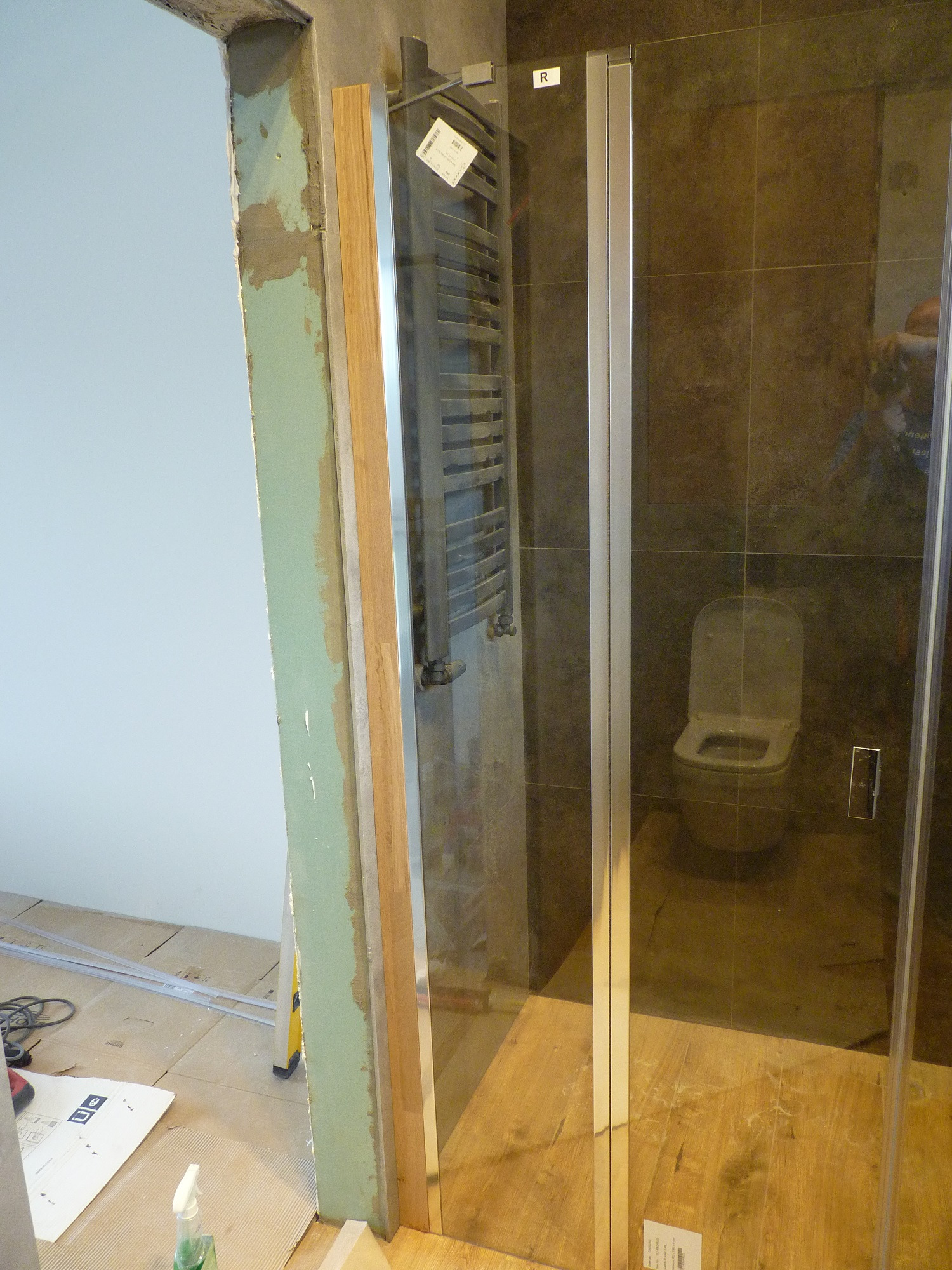 Łazienka w trakcie remontu: widoczna kabina prysznicowa z chromowanymi profilami, surowa ściana z resztkami zaprawy, grzejnik drabinkowy i częściowo ułożona podłoga z paneli.