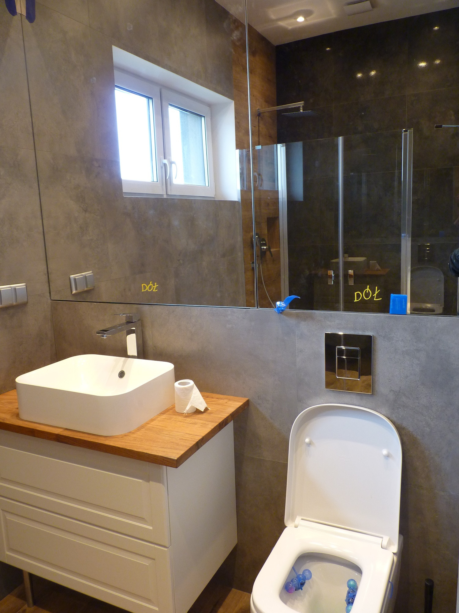 Nowoczesna łazienka z umywalką nablatową na drewnianym blacie, wiszącą toaletą z chromowanym przyciskiem spłukiwania i szklaną kabiną prysznicową w tle, na lustrze widoczne napisy 'Dół'