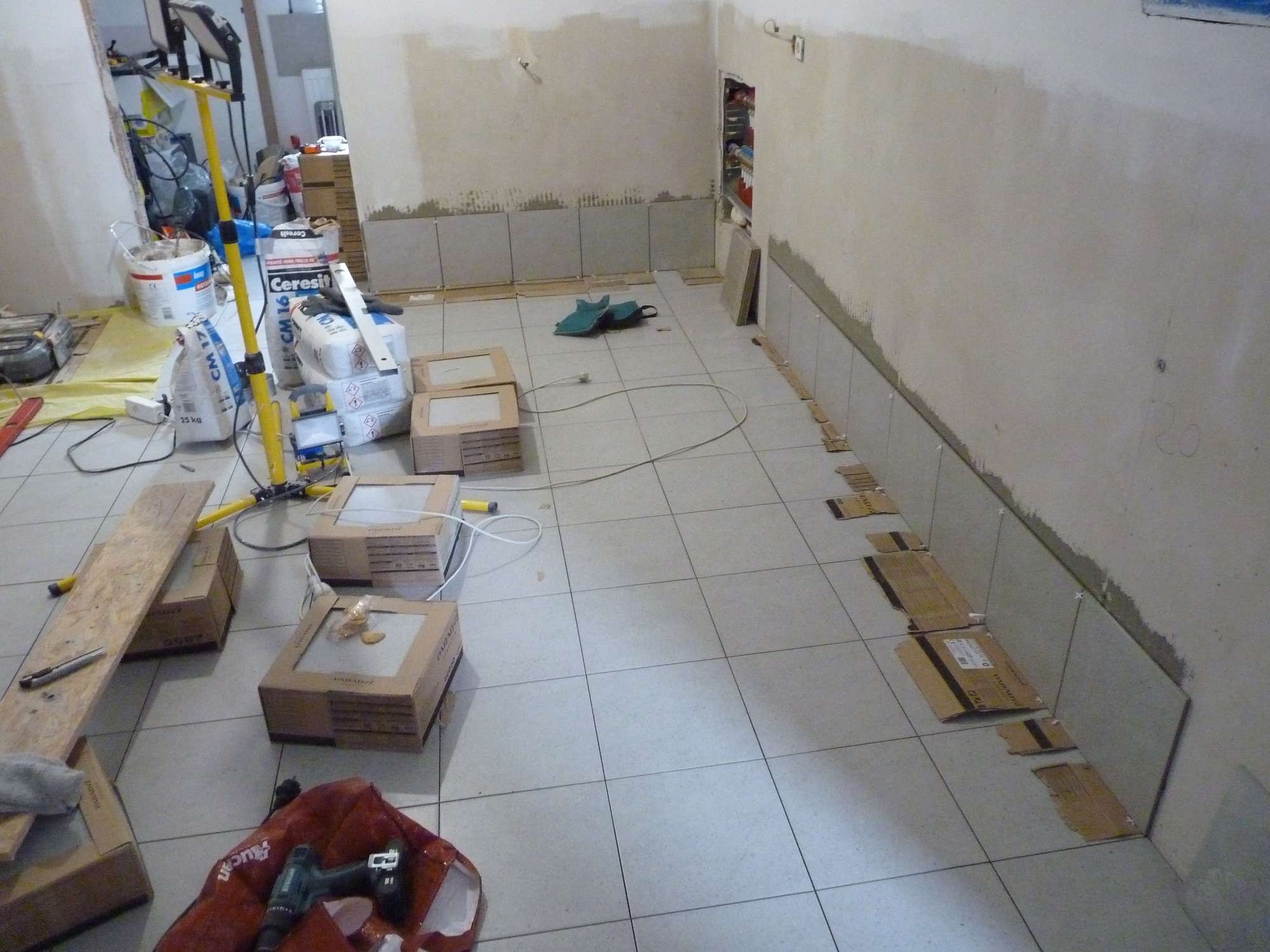 Przygotowania do układania płytek w łazience: częściowo wyłożone płytki na ścianie, paczki płytek na podłodze, narzędzia i worki z zaprawą w tle, widoczny fragment korytarza.