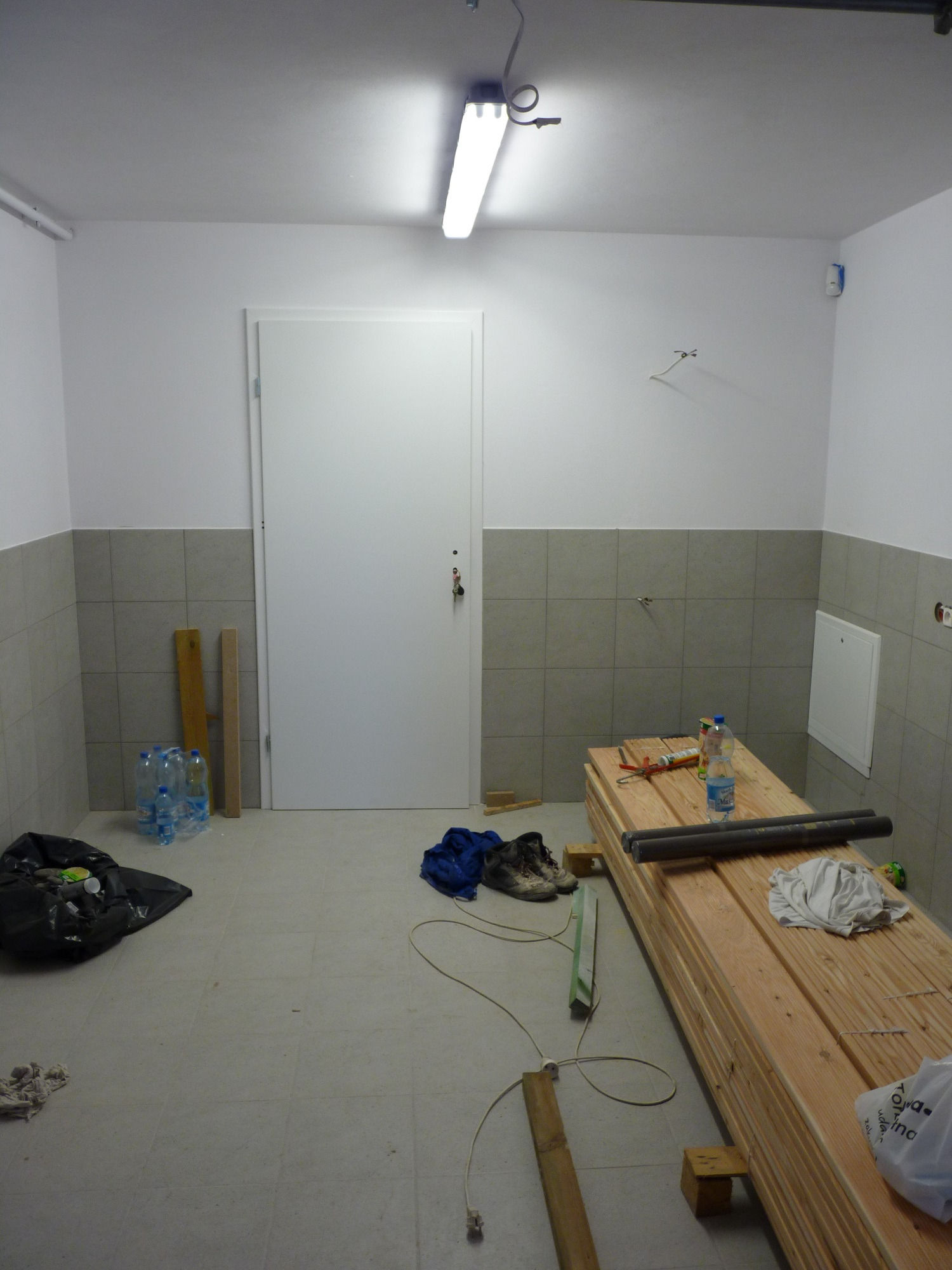 Pomieszczenie w trakcie prac wykończeniowych: ułożone do połowy płytki, stos desek, narzędzia, butelki z wodą i worek na śmieci.