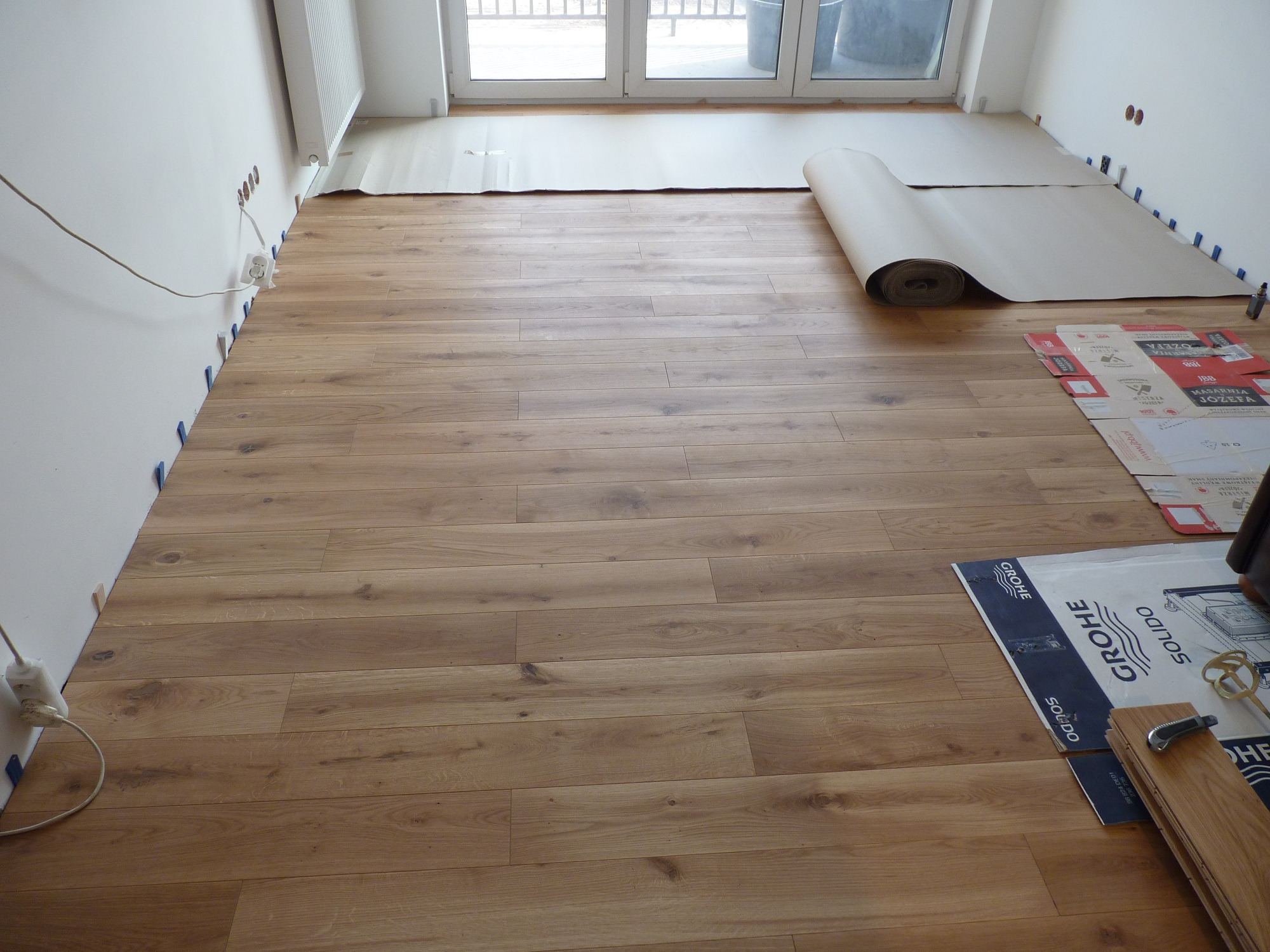 Układanie parkietu w jasnym pomieszczeniu, widoczne deski, podkład z rolki, materiały budowlane i narzędzia przy ścianie, niebieskie kliny dystansowe przy ścianie.