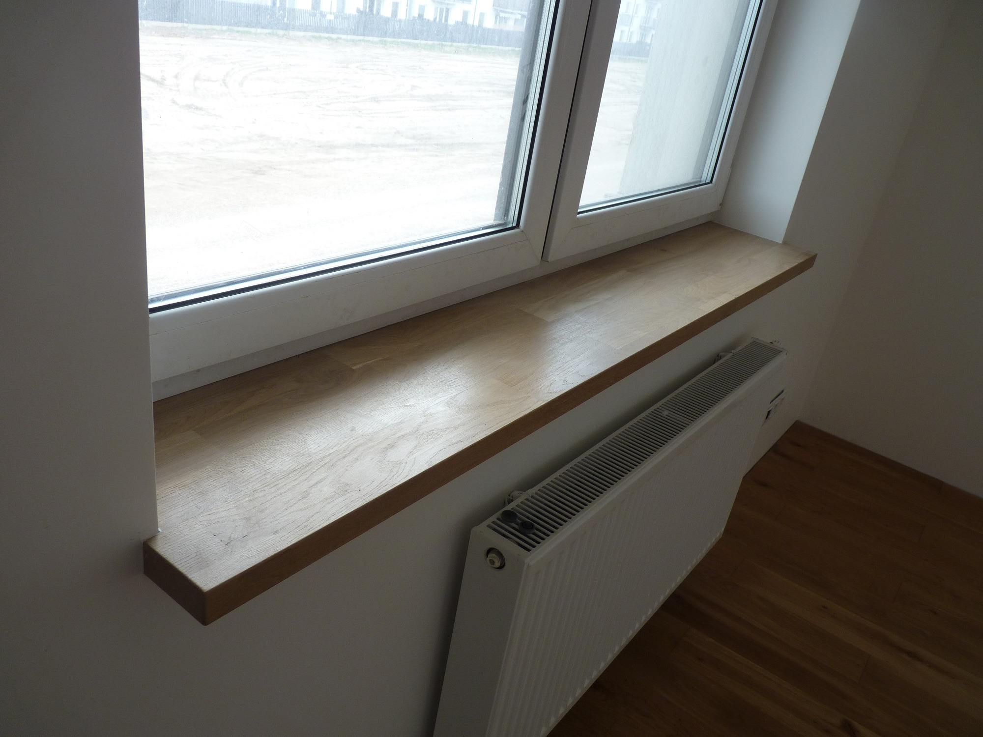 Jasny, drewniany parapet zamontowany pod białym oknem PCV, z widocznym grzejnikiem i fragmentem drewnianej podłogi w tle.