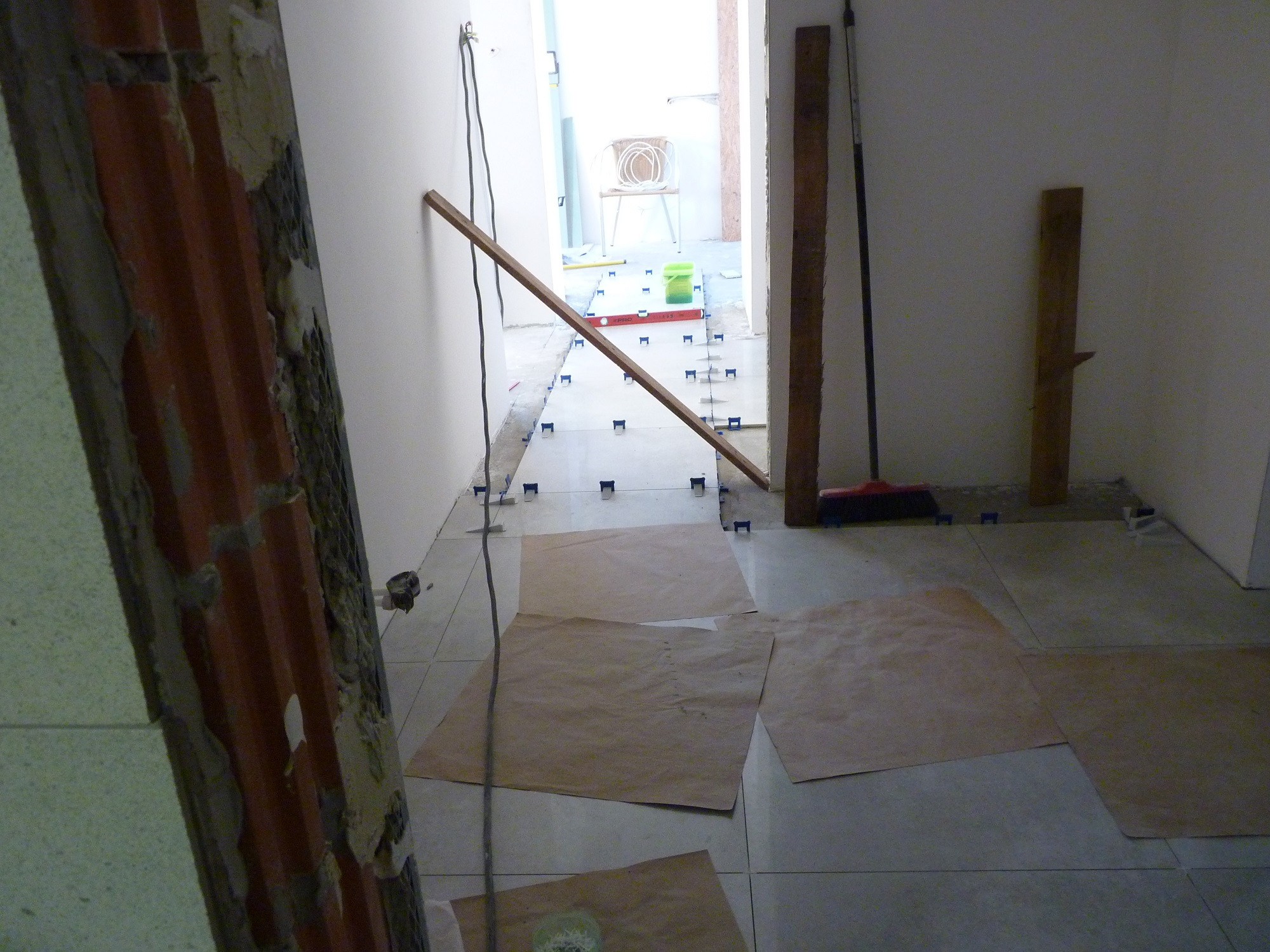 Układanie płytek podłogowych w korytarzu, widoczne płytki, poziomica, sznurek traserski, dystanse do płytek, kartony ochronne na podłodze.