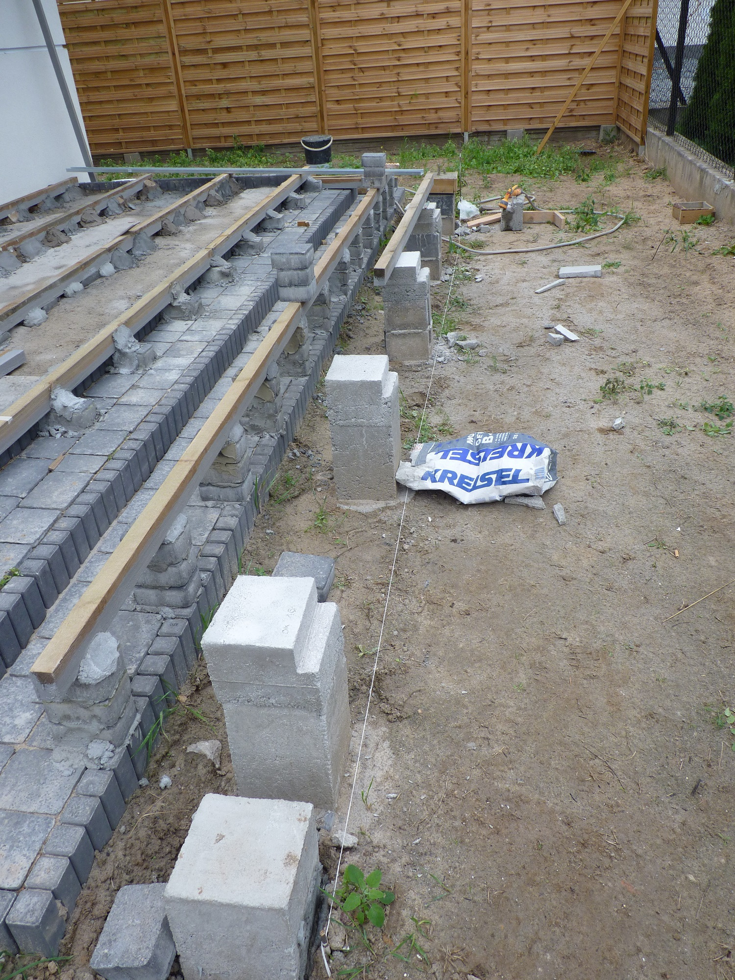 Budowa tarasu: betonowe bloczki fundamentowe, szara kostka brukowa i drewniane legary tworzą szkielet pod przyszłą konstrukcję, widoczny worek po cemencie Kreisel.