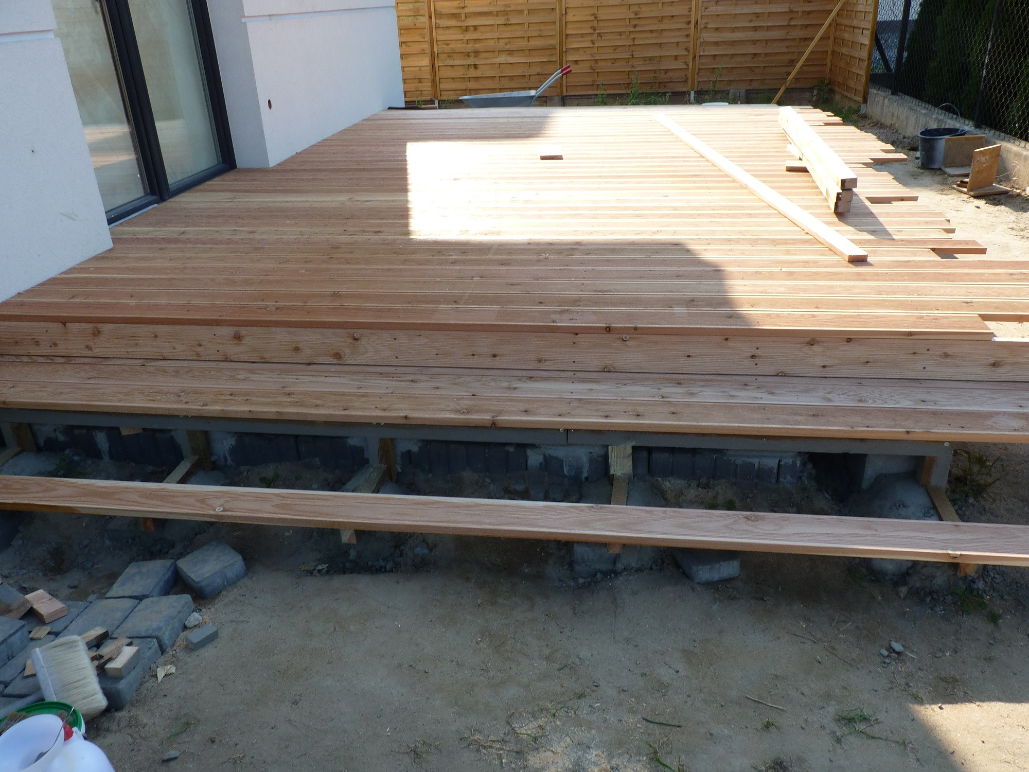 Budowa drewnianego tarasu: deski ułożone na konstrukcji, widoczne fundamenty, kostka brukowa i narzędzia w tle, częściowo zacienione