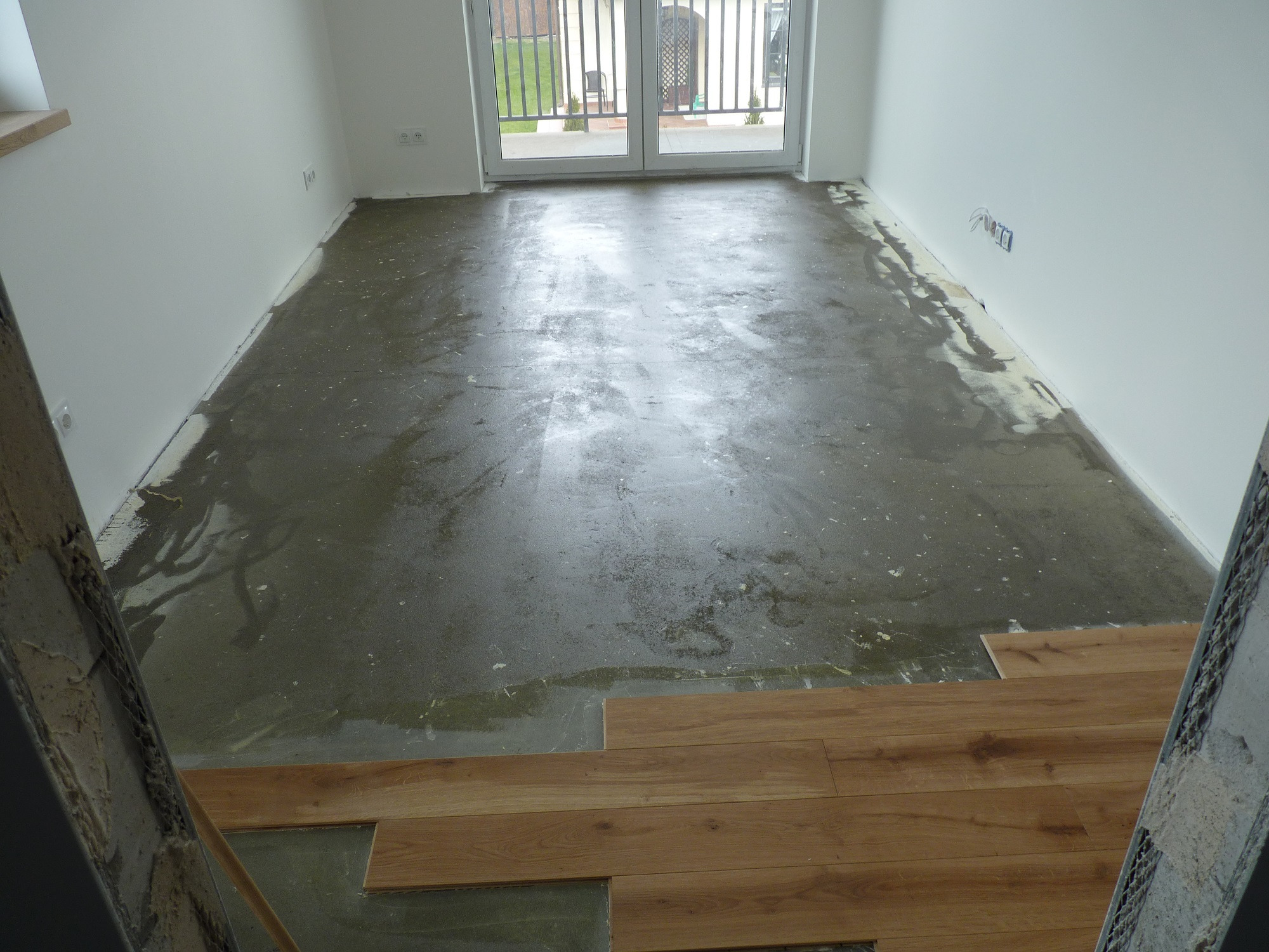 Przygotowanie podłoża pod panele podłogowe: wylewka wyrównująca z widocznymi śladami po szpachlowaniu oraz fragment ułożonych paneli w jasnym odcieniu drewna, widok z perspektywy wejścia...