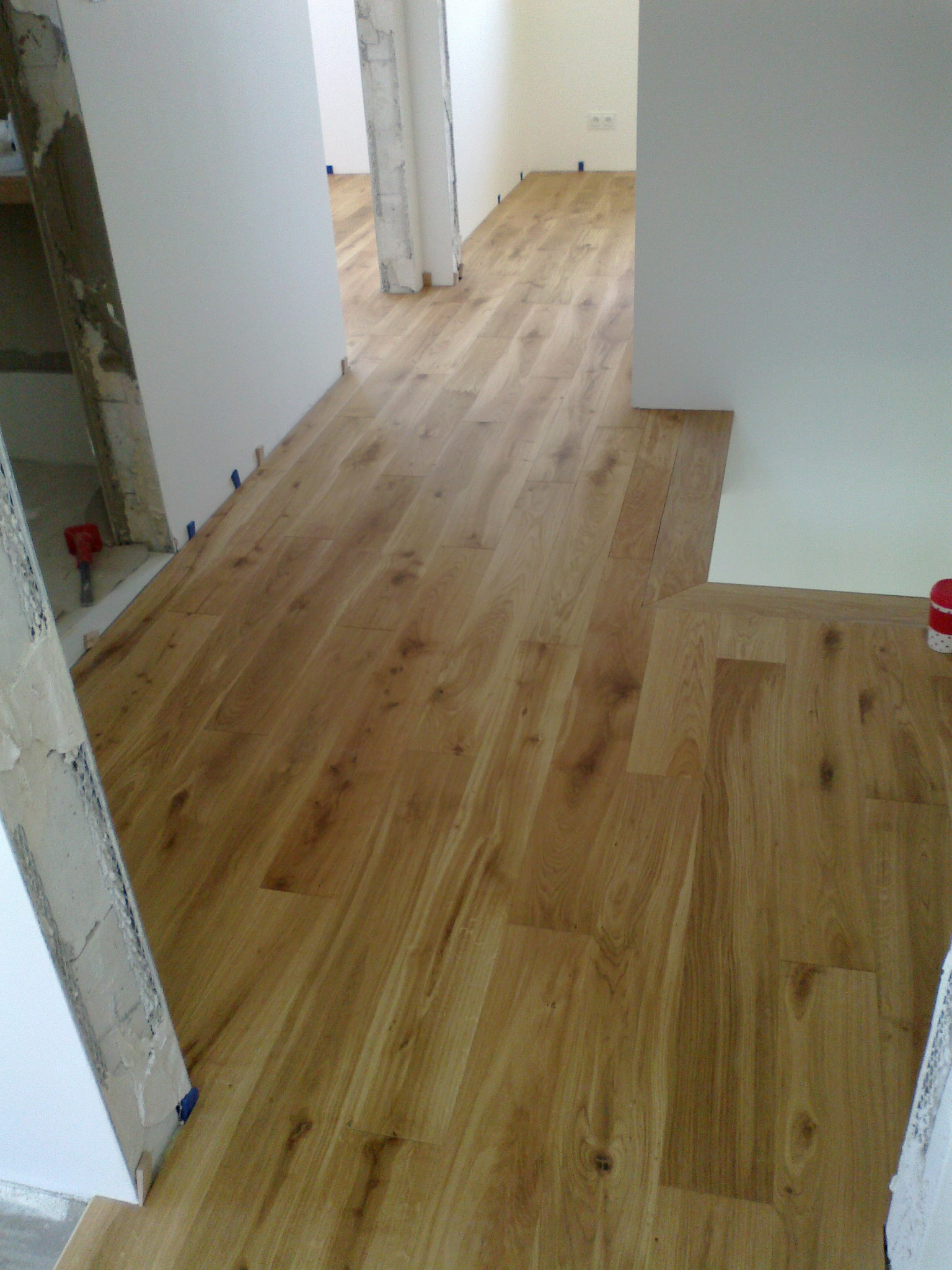 Fragment pomieszczenia z ułożoną podłogą z paneli drewnopodobnych, widoczne niedokończone wykończenia ścian i przejść, niebieska taśma maskująca przy listwach przypodłogowych.