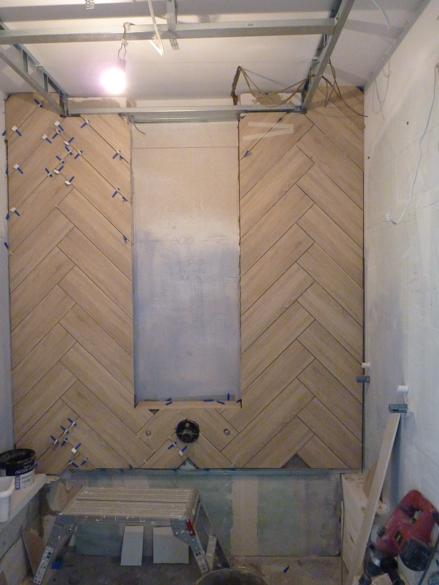 Montaż płytek imitujących drewno w jodełkę na ścianie w łazience, widoczne krzyżyki dystansowe, fragment sufitu z metalowym stelażem, niezabudowana wnęka i drabinka aluminiowa.