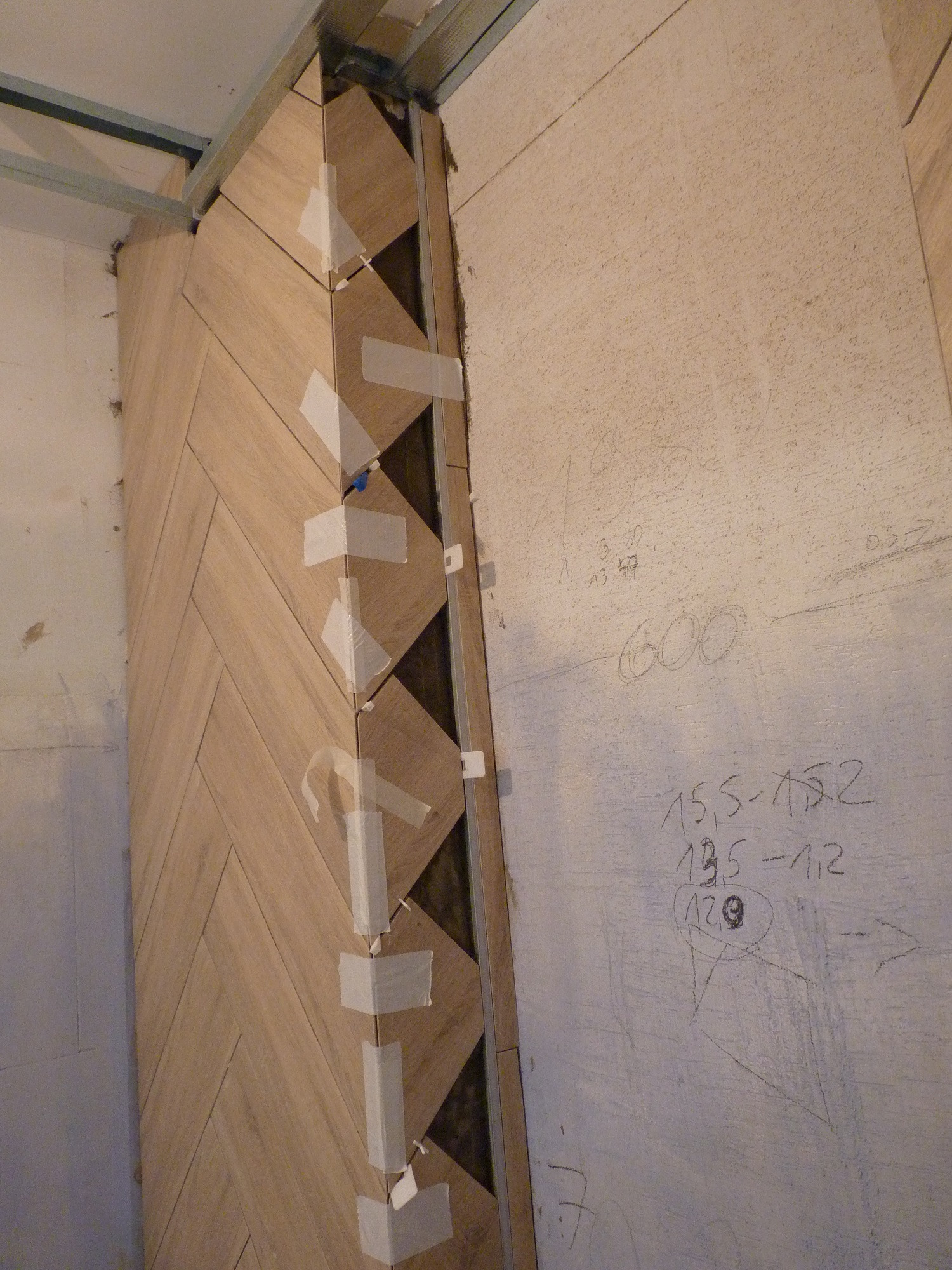 Montaż płytek imitujących drewno w jodełkę na ścianie, zabezpieczonych taśmą malarską, widoczne profile montażowe i notatki ołówkiem na sąsiedniej ścianie.