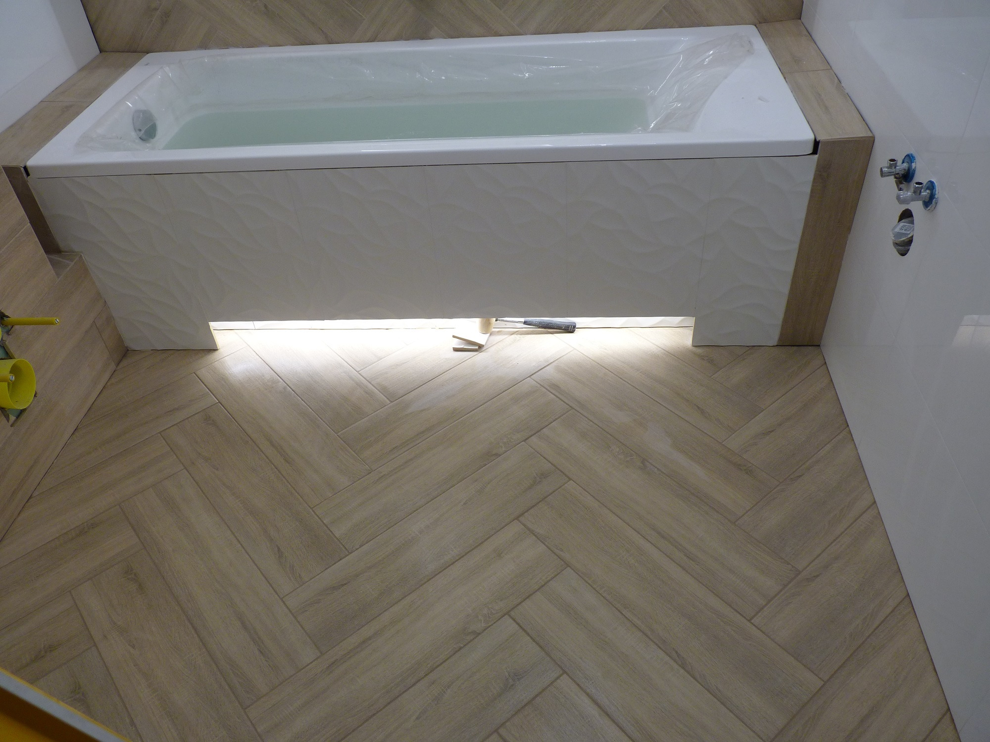 Nowa łazienka z wanną obudowaną płytkami o falistej strukturze, podświetleniem LED i podłogą z płytek imitujących drewno ułożonych w jodełkę; widoczne narzędzia glazurnicze.