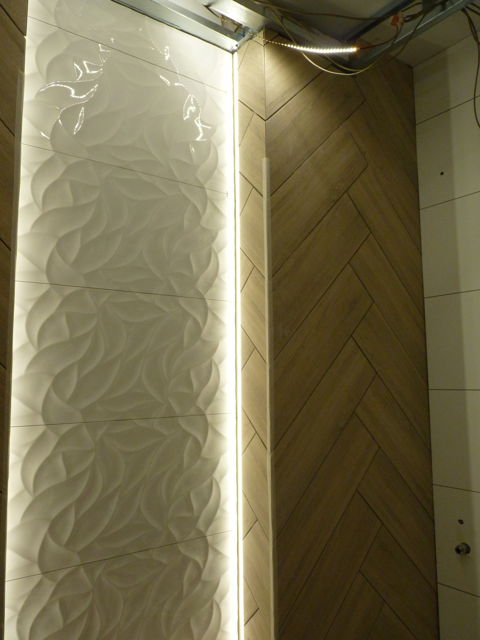 Fragment ściany łazienki z dekoracyjną, podświetloną pionową wstawką z białych płytek o falistym wzorze oraz drewnopodobnymi płytkami ułożonymi w jodełkę, widoczne kable i elementy montażowe...