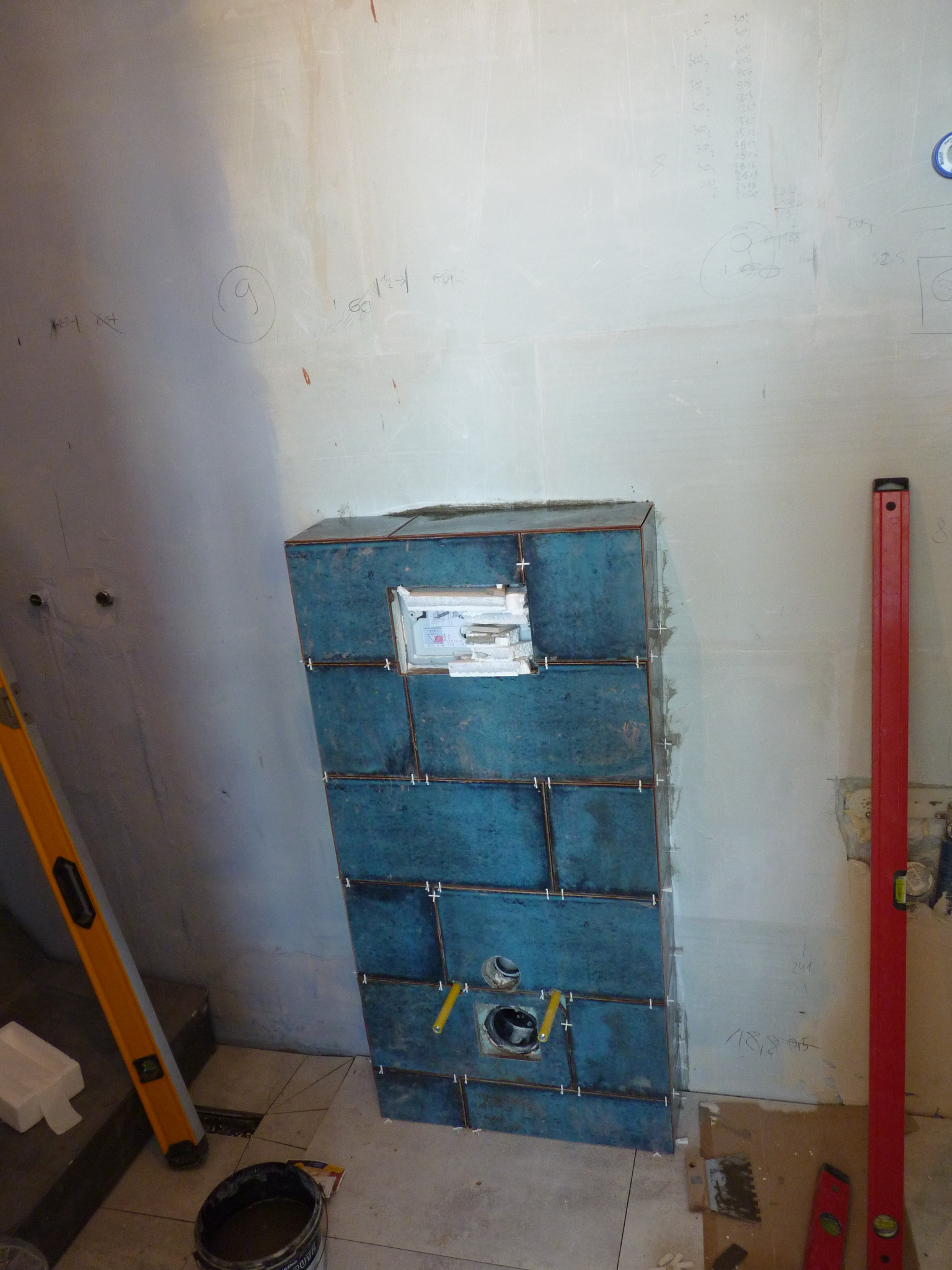 Montaż niebieskich płytek ceramicznych wokół podtynkowego stelaża WC w trakcie prac w łazience w Markach, widoczne poziomice i krzyżyki dystansowe.