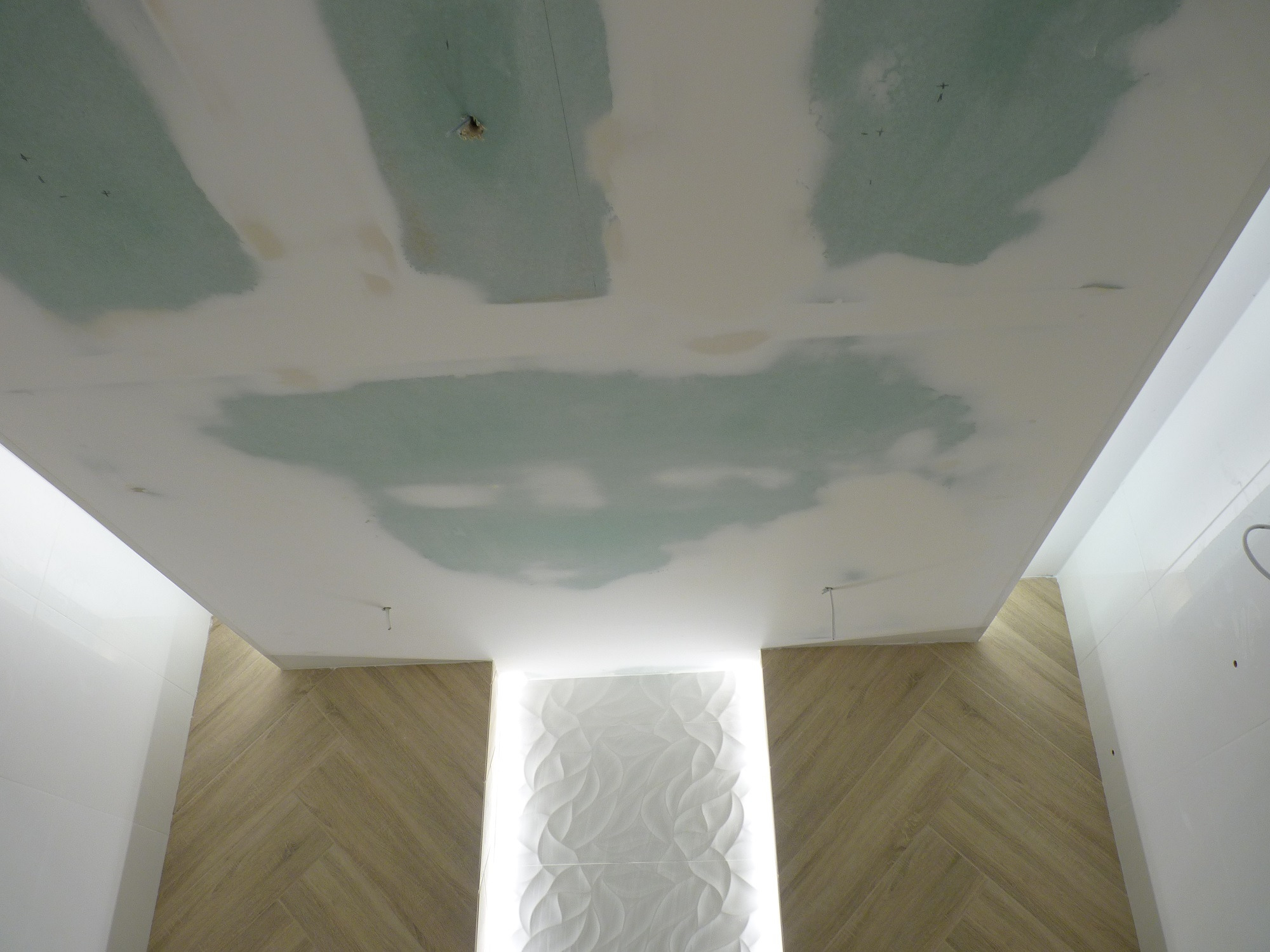 Niedokończony sufit z płyt kartonowo-gipsowych w łazience, widoczne ślady szpachlowania, oświetlona dekoracyjna ściana z trójwymiarowych paneli i płytki imitujące drewno ułożone w jodełkę.