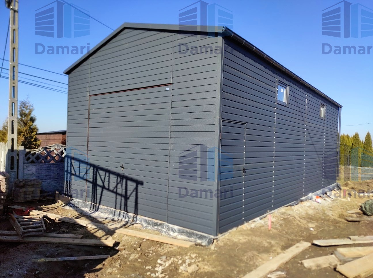 Nowoczesny, szary, dwuspadowy budynek gospodarczy z blachy trapezowej z małymi oknami, widoczny z boku, na tle błękitnego nieba i częściowo przygotowanego terenu.