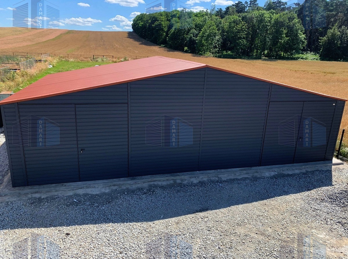 Duży, grafitowy budynek gospodarczy z czerwoną, metalową dachówką, widok z zewnątrz na tle pola i lasu.