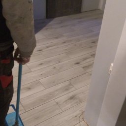 Dudulek - Czyszczenie nowo ułożonych paneli podłogowych mopem z niebieską nakładką, widoczne wystające kable elektryczne przy ścianie i szara, drewnopodobna faktura paneli.