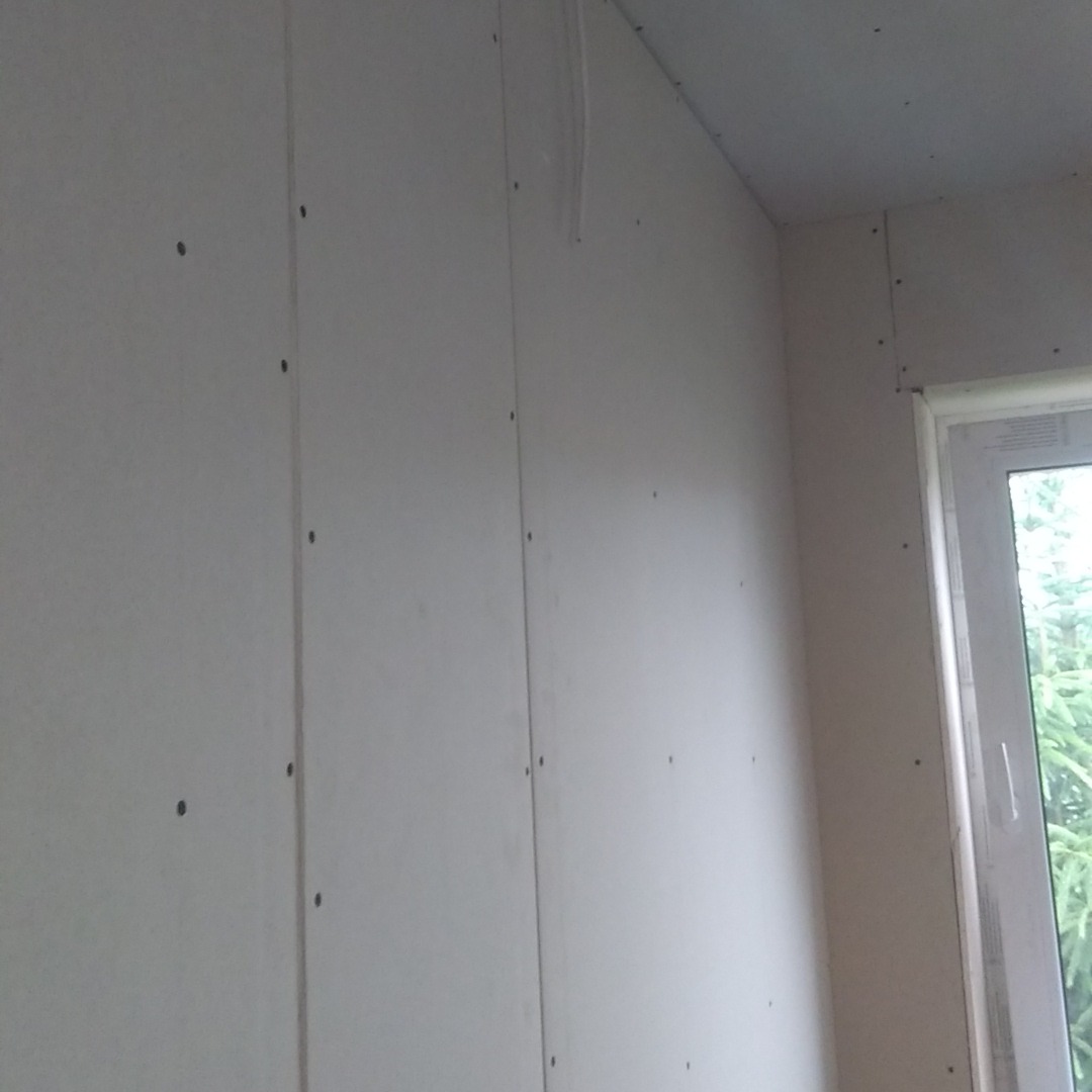 Ściana wykończona płytami kartonowo-gipsowymi, widoczne łączenia i wkręty, surowy stan przed szpachlowaniem, fragment okna w prawym górnym rogu.