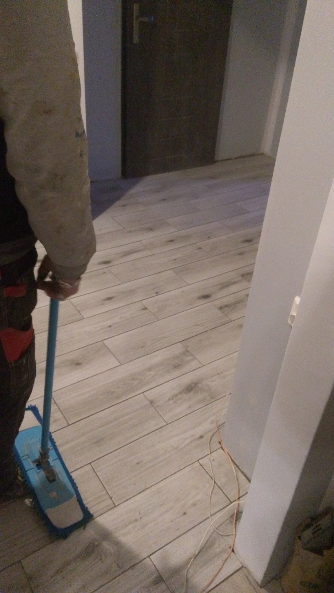 Czyszczenie nowo ułożonych paneli podłogowych mopem z niebieską nakładką, widoczne wystające kable elektryczne przy ścianie i szara, drewnopodobna faktura paneli.