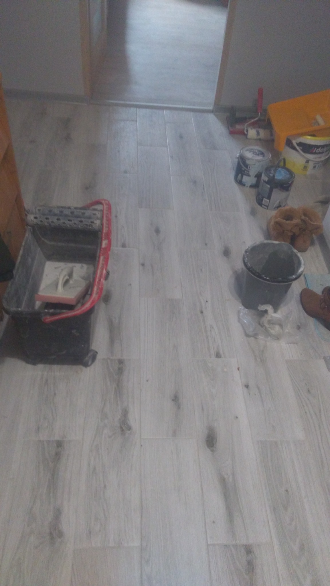 Jasne panele podłogowe z widocznym wiaderkiem z zaprawą, puszkami farb i narzędziami malarskimi w tle, wskazującymi na prace wykończeniowe.