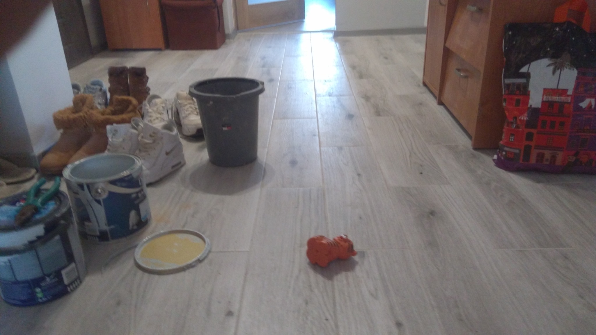 Podłoga z szarych paneli z widocznymi śladami po remoncie: puszki z farbą, pędzel, wiadro, buty robocze i zabawka w kształcie tygrysa
