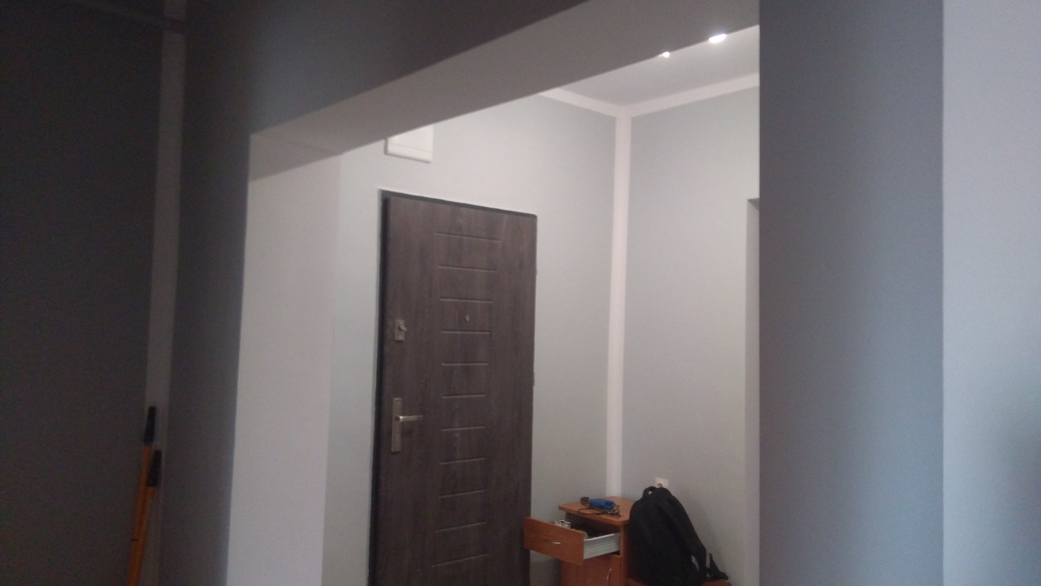Szare wnętrze mieszkania z ciemnymi drzwiami wejściowymi i meblami w tle, widok zza ściany.