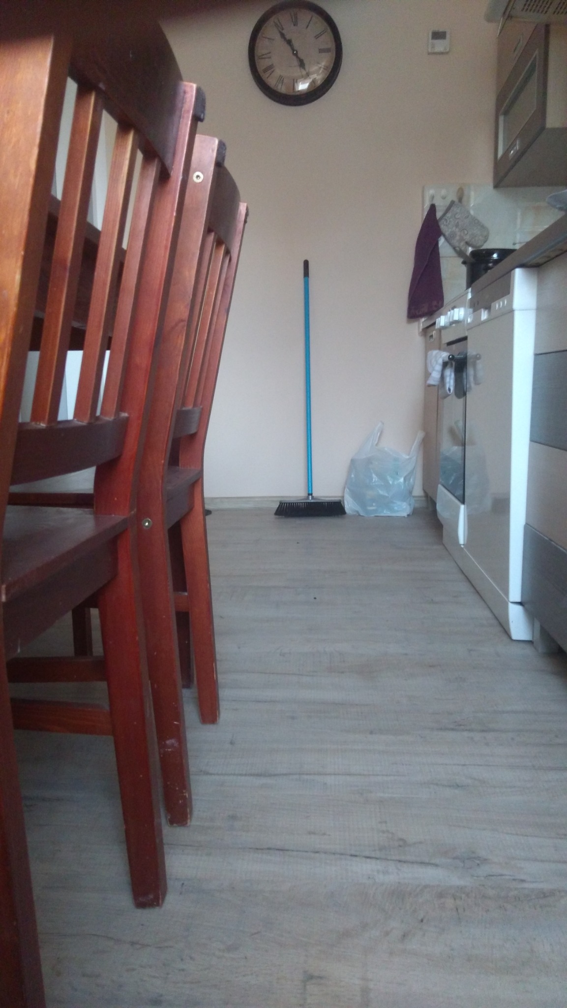 Fragment kuchni z widocznymi panelami podłogowymi, krzesłami z drewna, niebieską zmiotką i plastikową torbą na podłodze, zegar na ścianie.