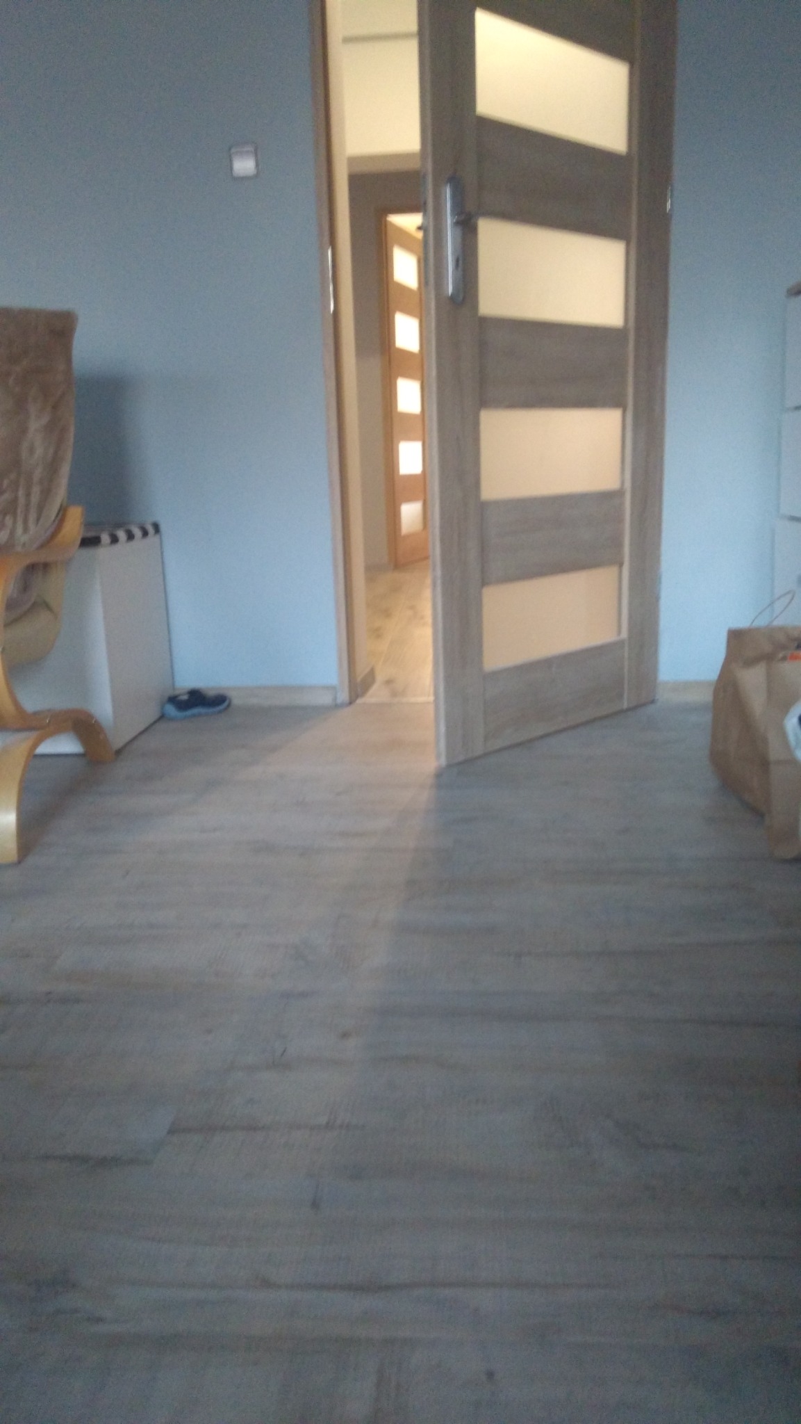 Jasne panele podłogowe ułożone w pokoju z szarą ścianą, widoczne otwarte drzwi wewnętrzne z przeszkleniami i fragment krzesła.