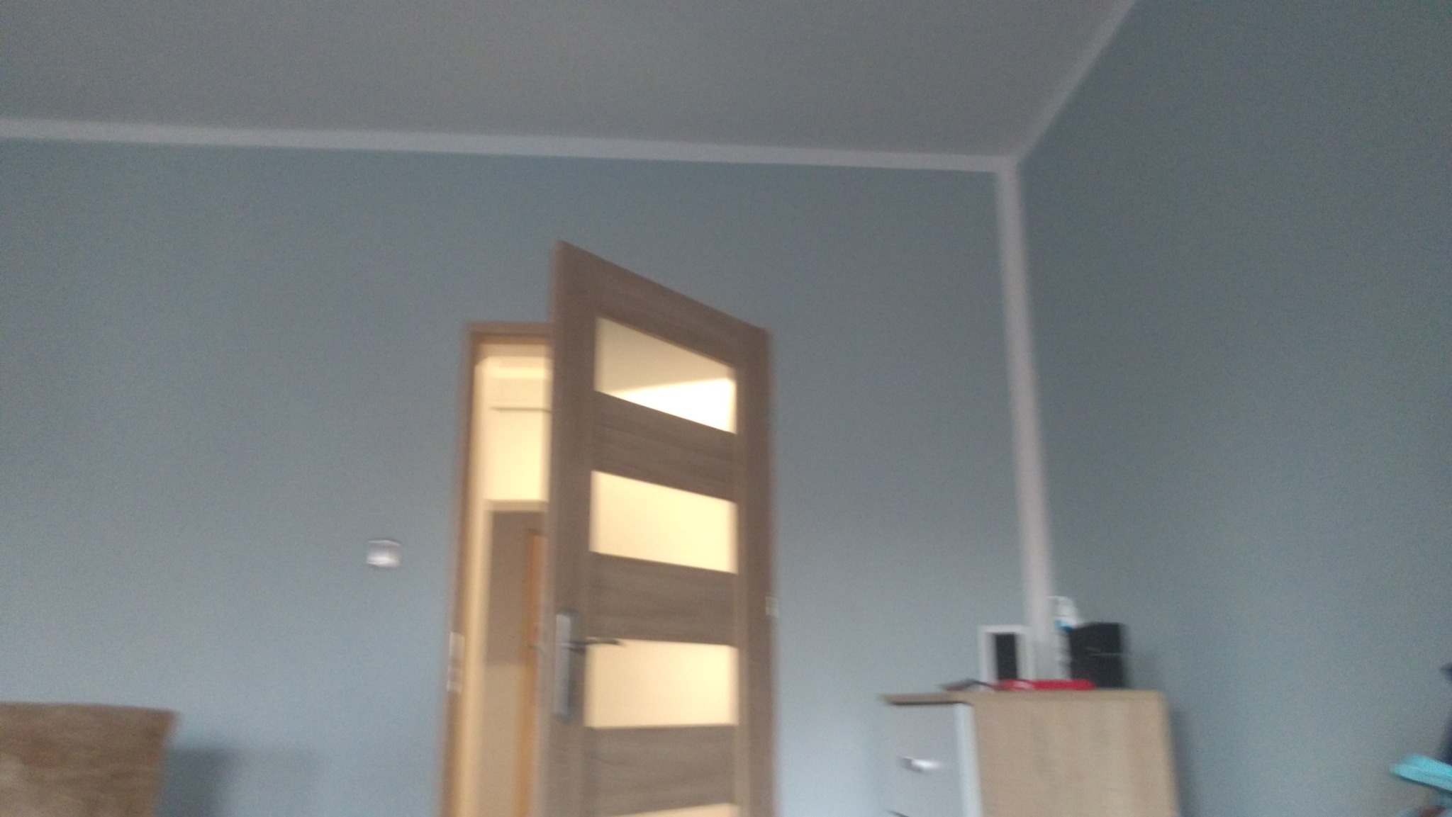 Otwarta, nowoczesna, szara, wewnętrzna stolarka drzwiowa z czterema poziomymi przeszkleniami, z widocznym fragmentem drugich drzwi w tle, na tle błękitnej ściany i białego sufitu.