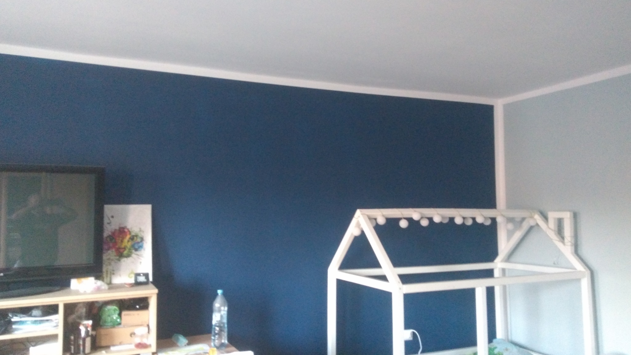 Pomieszczenie po malowaniu ścian na kolor granatowy i jasnoniebieski, widoczny biały sufit i listwa przypodłogowa, a także rama łóżka dziecięcego w kształcie domku z lampkami.
