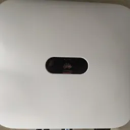 Biały inwerter Huawei zamontowany na szarej ścianie, widoczne kable w dolnej części urządzenia.