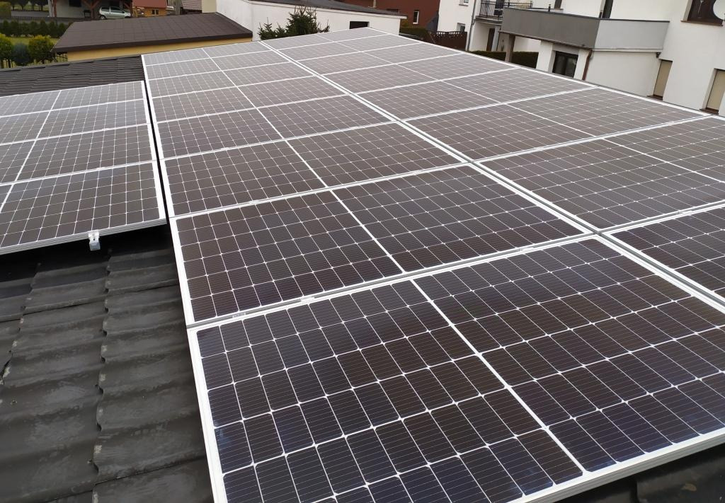 Panele słoneczne zamontowane na dachu domu jednorodzinnego z widocznymi elementami mocującymi i otaczającą zabudową.