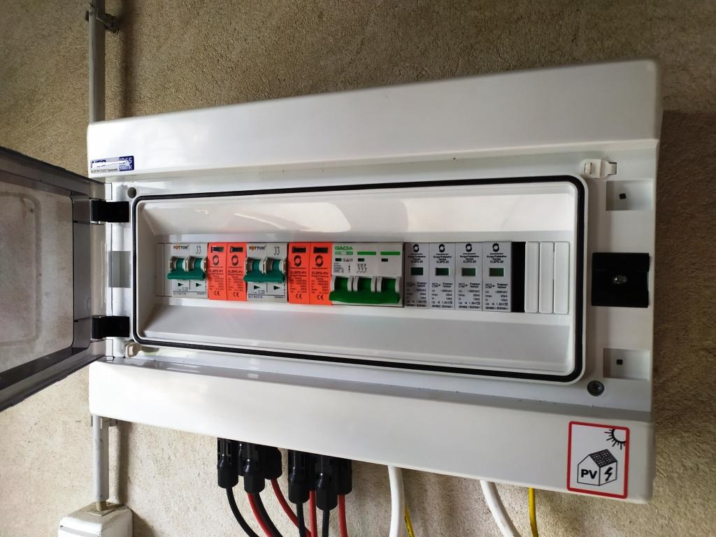 Rozdzielnica elektryczna zabezpieczeń DC instalacji fotowoltaicznej, z otwartymi drzwiczkami, widoczne bezpieczniki i ograniczniki przepięć, złącza kablowe na dole obudowy, naklejka ostrzegawcza PV.