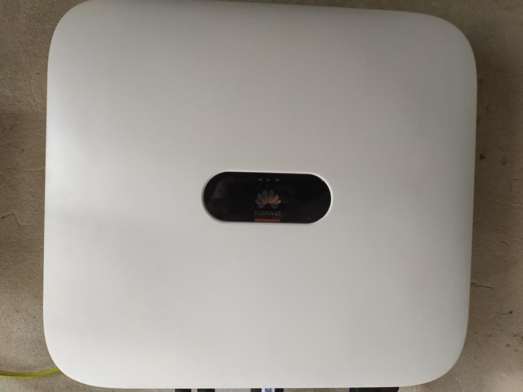 Biały inwerter Huawei zamontowany na szarej ścianie, widoczne kable w dolnej części urządzenia.