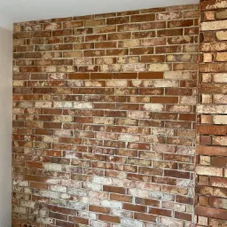 Ściana z dekoracyjnej cegły w odcieniach czerwieni, brązu i bieli, z widocznymi fugami i fragmentem narożnika. Na dole widoczne otwory instalacyjne w ścianie.