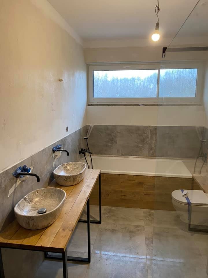 Nowoczesna łazienka w trakcie wykańczania: dwie kamienne umywalki na drewnianym blacie z metalowymi nogami, wanna obudowana drewnopodobnymi płytkami, szare płytki na ścianach i podłodze, widoczne...