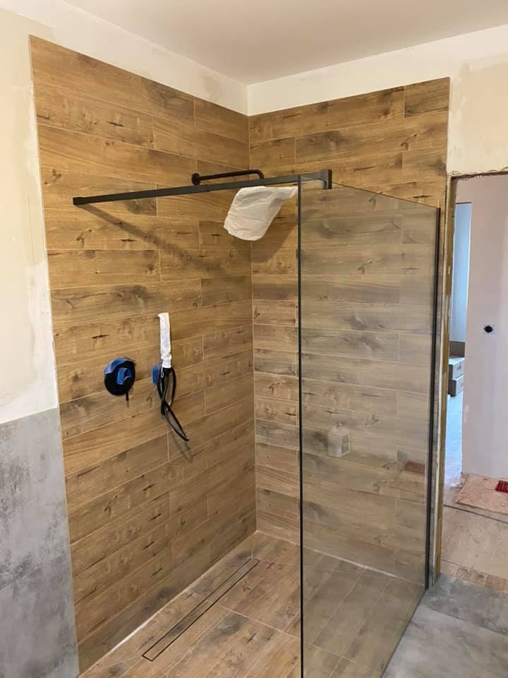 Montaż kabiny prysznicowej z drewnopodobnymi płytkami, widoczne przyłącza wodne i odpływ liniowy w trakcie prac wykończeniowych, zabezpieczona armatura.