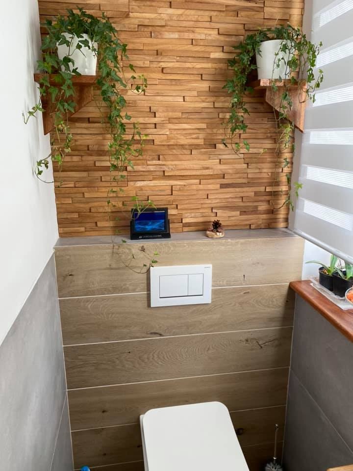 Nowoczesna łazienka z drewnianą mozaiką na ścianie, wiszącymi półkami z roślinami, drewnopodobnymi płytkami, białym sedesem i przyciskiem spłukującym Geberit.