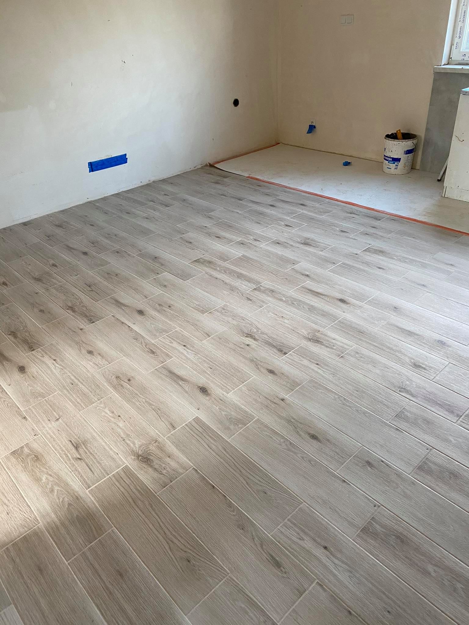Ułożona pod kątem podłoga z płytek imitujących drewno w jasnym odcieniu, widoczne fragmenty ścian z niezagruntowaną powierzchnią i niebieską taśmą malarską oraz wiadro z resztkami zaprawy.