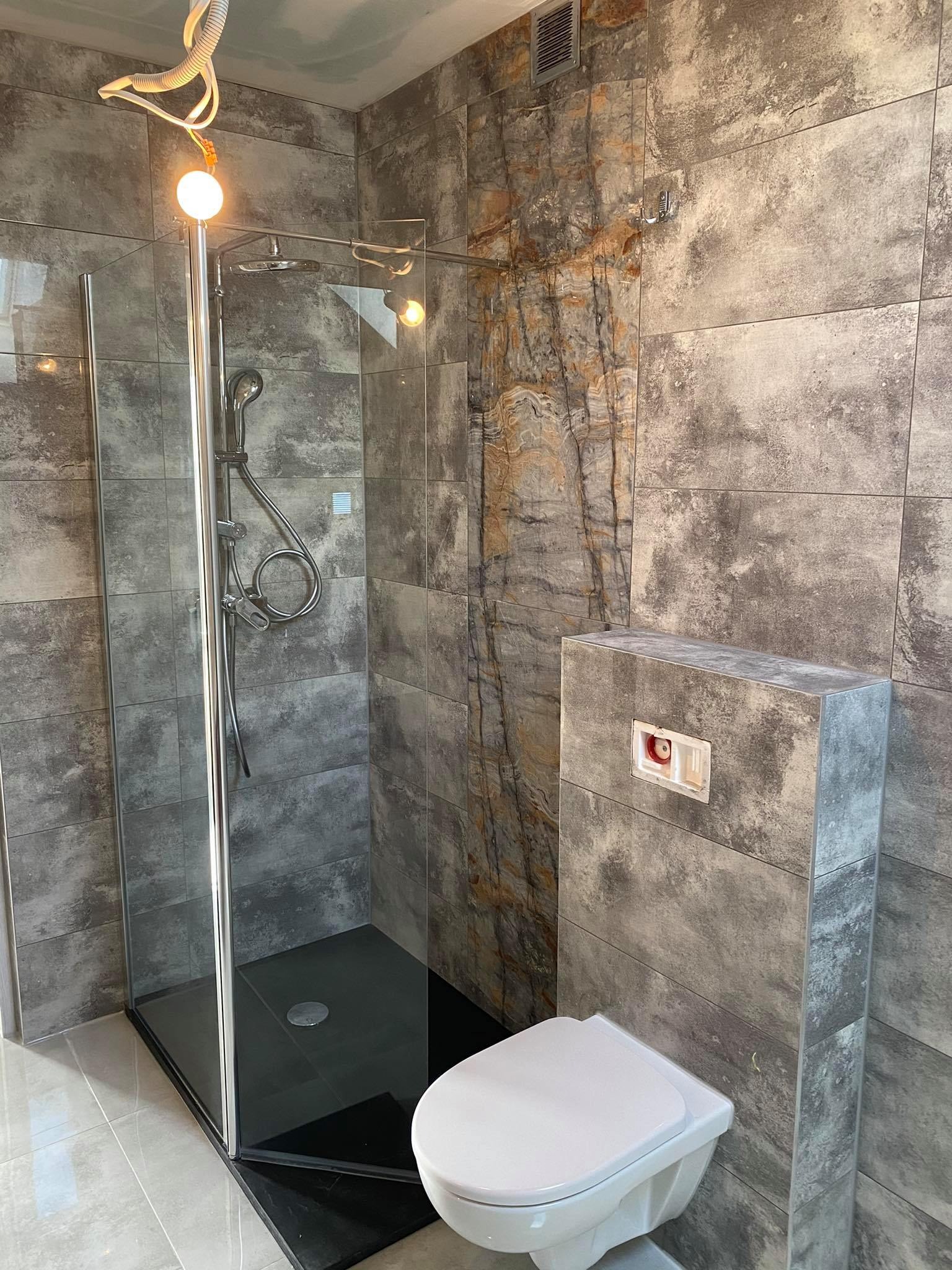 Nowoczesna łazienka z szarymi płytkami imitującymi kamień, kabiną prysznicową z czarnym brodzikiem i podwieszaną toaletą.