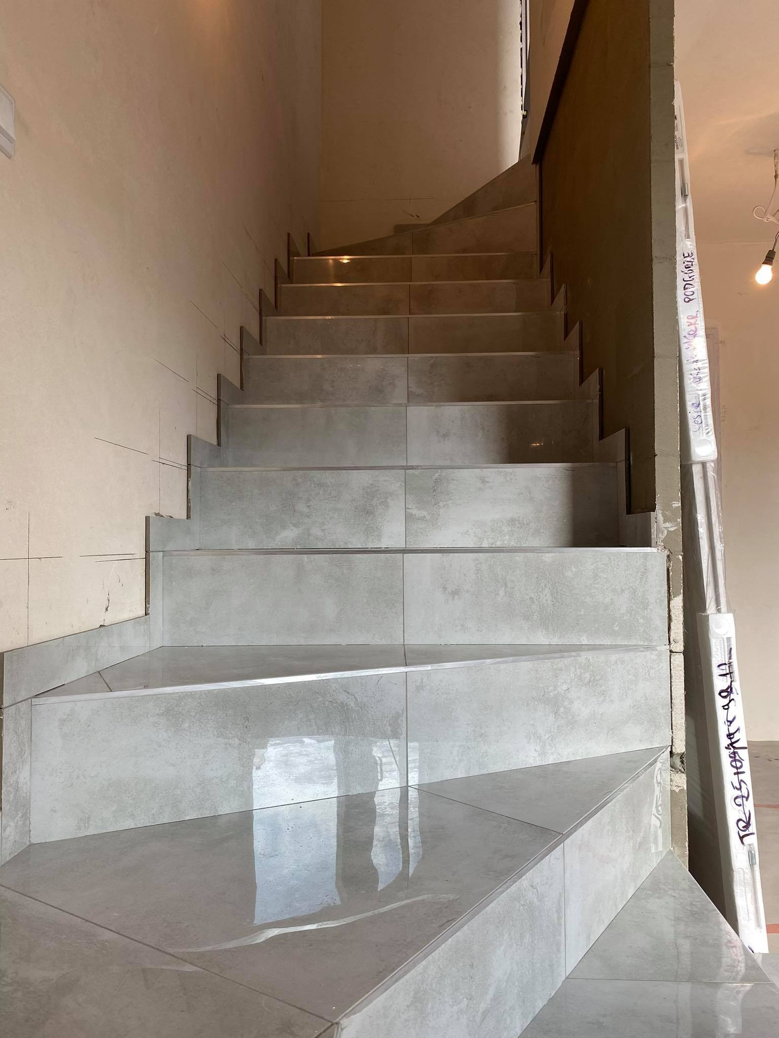 Wykończone schody wewnątrz budynku, pokryte szarymi płytkami ceramicznymi o nieregularnym kształcie, widoczne ślady po ołówku na ścianie, wskazujące na etap prac wykończeniowych.