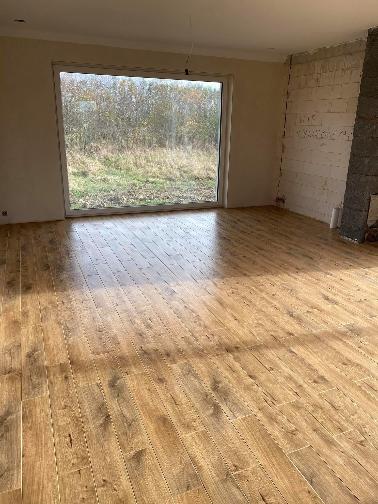 Ułożona podłoga z paneli imitujących drewno w surowym pokoju z dużym oknem i ścianami w stanie deweloperskim.