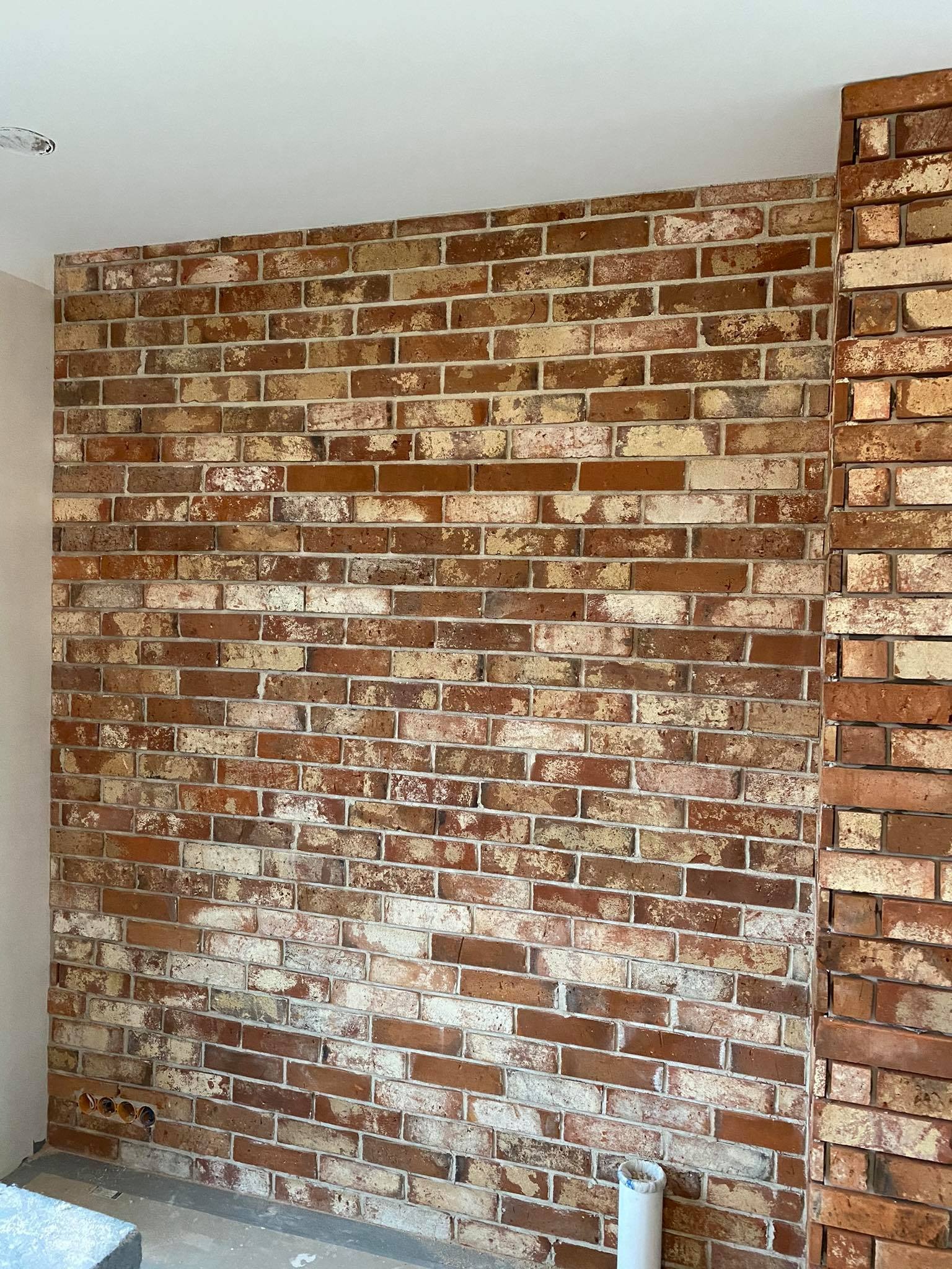 Ściana z dekoracyjnej cegły w odcieniach czerwieni, brązu i bieli, z widocznymi fugami i fragmentem narożnika. Na dole widoczne otwory instalacyjne w ścianie.