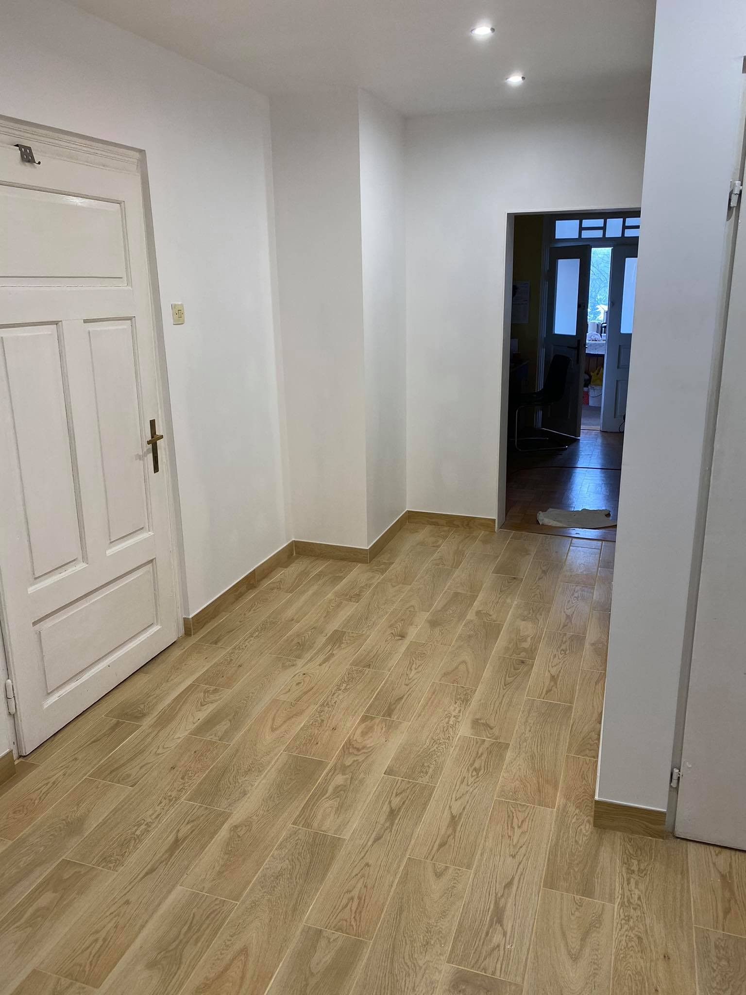 Korytarz z nową podłogą imitującą drewno, białe ściany i drzwi, widok do pokoju z oknem balkonowym.