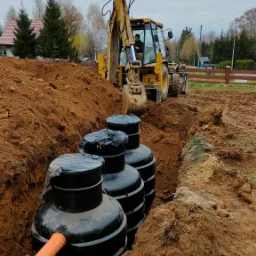 Montaż czarnych zbiorników na szambo w wykopie ziemnym, widoczny pomarańczowy rurociąg i operator koparki w żółtej maszynie budowlanej w tle.