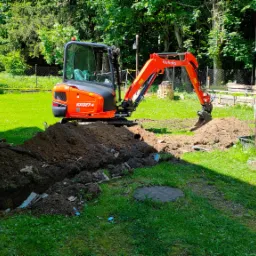 Pomarańczowa minikoparka Kubota KX027-4 wykonuje wykop w trawiastym terenie obok drewnianego budynku, widoczne ułożone kostki brukowe i zwój niebieskiego węża.